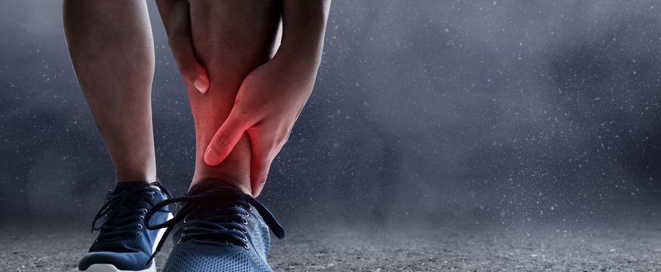 person mit dunkelblauen laufschuhen hält sich den rot markierten knöchel vor schmerz vor dunklem hintergrund