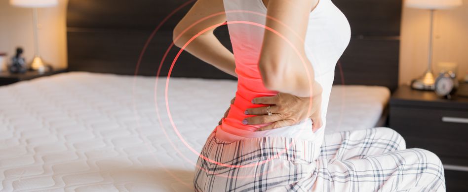 frau sitzt auf bett und hält sich den rücken vor schmerz an rot markierter stelle