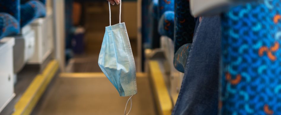 frau hält medizinische maske an bändeln in bahn mit überschlagenen beinen jeans schwarzem pulli und weißen schuhen