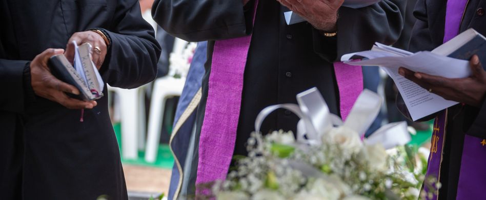 pfarrer steht vor sarg mit lila schwarzem anzug und hält trauerrede