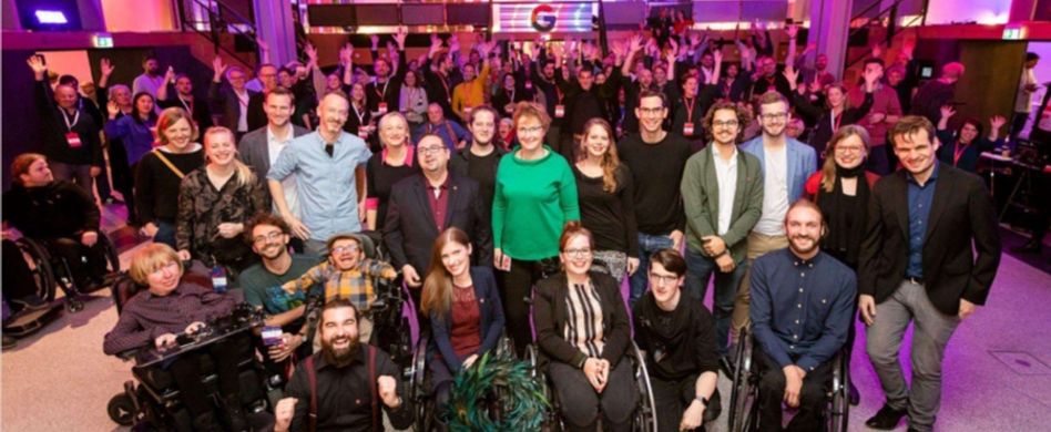 Eine große Gruppe von Menschen mit und ohne Behinderungen positioniert sich in einem Tanzsaal zu einem geselligen Gruppenbild.