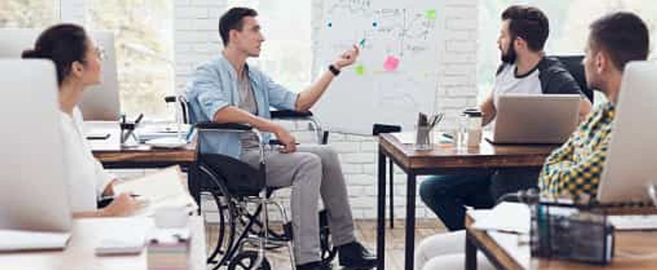 Ein Mann im Rollstuhl hält eine Präsentation in einem Büro vor mehreren Personen.