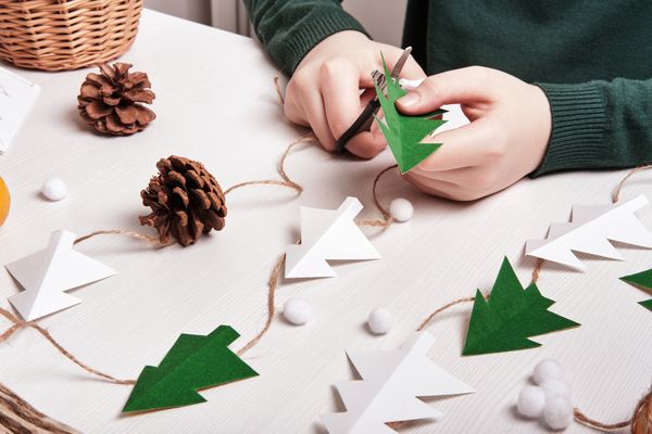 kind bastelt weihnachtsbäume aus papier mit tannenzapfen und kordel auf weißem hintergrund